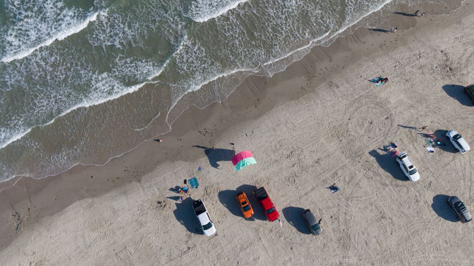 Top-down aerial shot of a sunny day at the beach in Corpus Christi, Texas with cars parked along the shoreline.