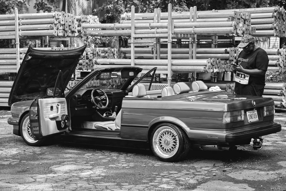 A man inspects a vintage convertible car in an industrial outdoor area.